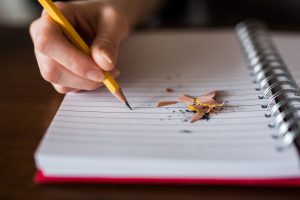 imagem de uma mão escrevendo à lápis em um caderno em branco com raspas de lápis apontado